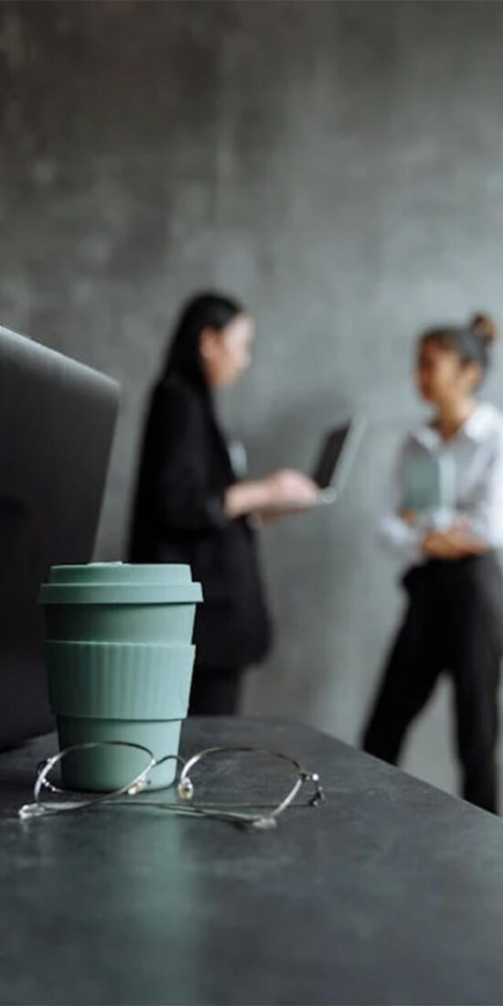 Laptop, coffee cup, and glasses on a desk with two professionals discussing business strategies in a modern office, symbolizing business development.