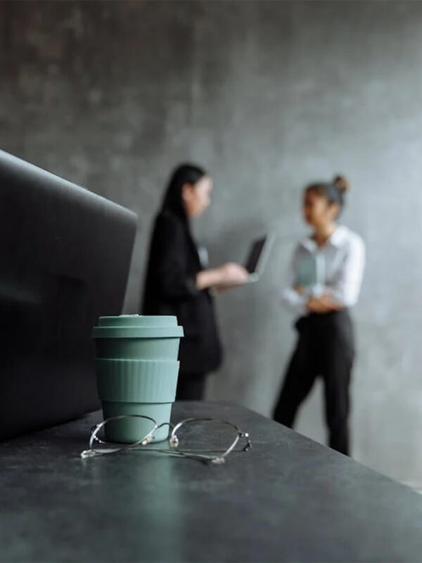 Laptop, coffee cup, and glasses on a desk with two professionals discussing business strategies in a modern office, symbolizing business development.