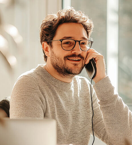Smiling man in glasses and a sweater talking on the phone in a bright office setting, representing Robert Fox, Founder & CEO of a consulting firm focused on analytics-driven solutions and team engagement.